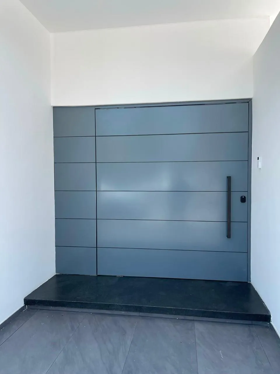 Puerta pivotante metálica gris en acceso principal de vivienda